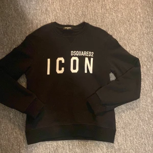 Svart sweatshirt DSQUARED2 ICON - Svart sweatshirt från DSQUARED2 med vit text 'ICON' och logga framtill. Klassisk rund halsringning och långa ärmar. Mjuk insida och ribbade muddar vid ärmslut och nederkant. Perfekt för en cool och avslappnad stil.