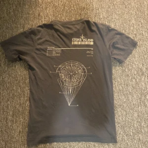 Svart Stone Island t-shirt med tryck - Svart t-shirt från Stone Island med vit logga på bröstet och stort grafiskt tryck på ryggen. Klassisk rund halsringning och korta ärmar. Tillverkad i mjuk bomull som känns skön mot huden. Perfekt för dig som gillar streetwear och vill sticka ut lite extra.