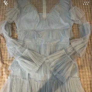 Ljusblå volangklänning med fransar - Superfin ljusblå klänning med v-ringning och långa ärmar. Klänningen har flera lager av volanger och fransdetaljer i ett skirt, tunt material som ger en riktigt drömmig känsla. Perfekt för dig som vill sticka ut med en unik och trendig look.