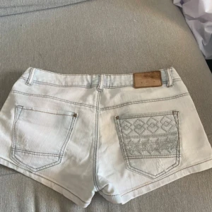 Vita jeansshorts från Lineen - Snygga vita jeansshorts från Lineen med normal midja och klassisk femficksdesign. Ena bakfickan har ett broderat mönster som ger en unik touch. Shortsen har ljus tvätt och är perfekta för varma dagar. Stängs med knapp och dragkedja.