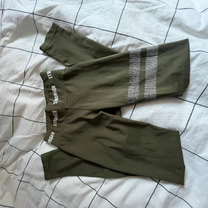 gröna tränings taights från Stronger - Snygga gröna träningsbyxor från Stronger med vita logotyper i midjan och två vita randiga detaljer på ena benet. Dem sitter taight och har hög midja, säljer för att dem är försmå, har används ganska många gånger och har slitits på de två vita detaljerna på benet, lite uttöjda på baksidan annars är dem i bra skick