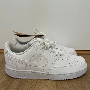 Vita sneakers från Nike , storlek 38 - Snygga helvita sneakers från Nike i storlek 38. Skorna har klassisk design med rund tå, snörning och platt sula. Tillverkade i syntetmaterial med perforerade detaljer framtill för extra stilpoäng. Perfekta för dig som gillar en clean och fräsch look.