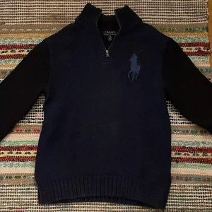 Mörkblå stickad tröja Polo Ralph Lauren - Mörkblå stickad tröja från Polo Ralph Lauren med halv dragkedja och hög krage. Stor broderad logga på bröstet.