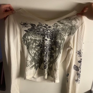 Vit långärmad topp  - Vit långärmad topp med svart tryck framtill och på ena ärmen. Tröjan har v-ringad hals och är ribbad i materialet, vilket ger en snygg struktur. Passformen är något croppad och tyget känns mjukt och stretchigt.