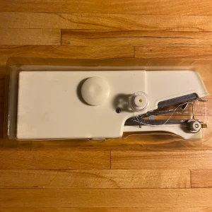 Hand Symaskin E-Z - Praktisk handhållen symaskin för snabba lagningar och fållningar. Perfekt för att laga kläder, korta byxor och sy gardiner. Enkel att använda, drivs av 4 AA-batterier (ej inkluderade). Inkluderar trådrulle och tillbehör. Oanvänd i originalförpackning.