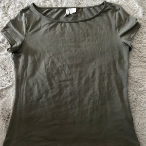 Olivgrön kortärmad topp från Divided - En basic olivgrön topp från Divided med rund halsringning och korta ärmar. Tillverkad i mjuk bomull, perfekt för en enkel och clean look. Passformen är normal och toppen är lätt att matcha med olika outfits.