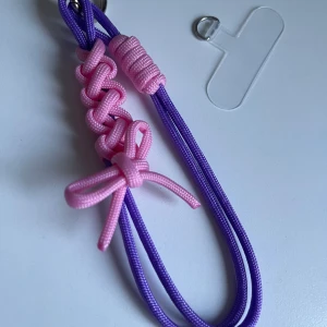 Lila och rosa handledsrem till mobil - Snygg handledsrem till mobilen i lila och rosa med flätad design och söt rosett. Perfekt accessoar för att göra din mobil mer personlig och lättare att bära med sig. Fästes enkelt i skalet med metallring. Gjord i slitstark syntet.
