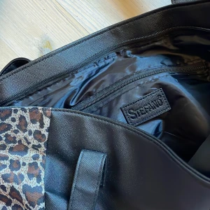 Svart väska med leoparddetaljer - Ursnygg väska från märket Stefano som passar till allt - både stunder och kläder. Väskan är rimlig med ett stort fack och två små (alla m dragkedjor som fungerar perfekt). Väskan är i ordentligt material och tål nog båda lite slit samt vatten. Väskan har jättesnyggt leopardmönster på sidorna som är där och gör väskan unik men ändå inte tar över. Älskar denna väska men kommer inte till användning alls. Sugen på en ny snygg skolväska? Eller bara för att spicea upp rummet eller outfiten? :)
