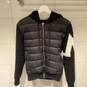 Svart M pufferjacka med huva - Säljer en svart pufferjacka från M med stickade ärmar och stor vit zigzagdetalj på ena ärmen. Jackan har huva, dragkedja framtill och snörning vid halsen. Perfekt för dig som vill ha en snygg och sportig look.