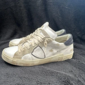 Philippe Model vita skor  - Snygga vita sneakers från Philippe Model med beige mockadetaljer och svart häl. Skorna har snörning, rund tå och en klassisk logotyp på sidan. Ovandelen är i skinn och mocka, med en platt sula i gummi. Perfekta för dig som gillar stilrena och trendiga sneakers.