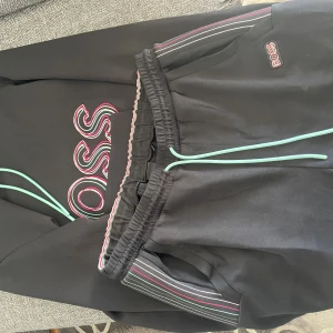 Svart hoodie från BOSS med färgglada detaljer - Svart dress från BOSS med stor broderad logga i rosa, vitt och grönt på bröstet. Tröjan har huva med mintgröna snören och långa ärmar. Snygga randiga detaljer i rosa och vitt på huva och ärmslut. Perfekt för en chill och trendig look.