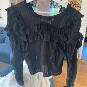 Svart blus med volanger - Säljer en svart blus med långa ärmar och volangdetaljer över bröstet och längs ärmarna. Blusen är lätt transparent med subtila ränder i tyget. Storlek M och i super bra skick 💕