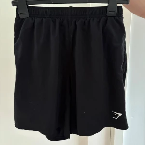 Svarta träningsshorts från Gymshark - Svarta shorts från Gymshark med elastisk midja och logga i vitt på benet, med loose passform och mjukt material som ger bra rörelsefrihet. Snygg och enkel design som funkar till alla träningspass.