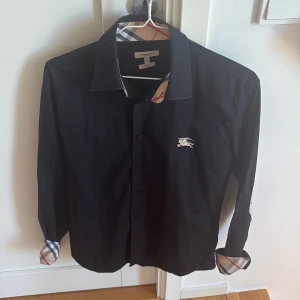 Svart Burberry skjorta slim fit M - Svart skjorta från Burberry London i slim fit med klassiskt rutigt mönster på insidan av krage, manschetter och armbågsdetaljer. Ikonisk Burberry-logga broderad på bröstet. Skjortan har långa ärmar och knappar framtill. Materialet är mjuk bomull.
