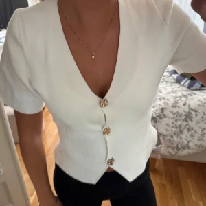 Vit kortärmad blus med guldknappar - Säljer en stilren vit blus med korta ärmar och djup v-ringning. Toppen har tre dekorativa guldknappar framtill och en snygg spetsig nederkant. Materialet känns mjukt och har en lätt stretch. 