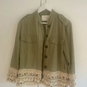 Grön overshirt från Zara med fransar - Snygg grön overshirt från Zara med unika virkade detaljer, fransar och guldiga knappar längs nederkanten och ärmsluten. Jackan har två bröstfickor, knäppning framtill och är tillverkad i bomull. Perfekt för dig som vill sticka ut med din stil.