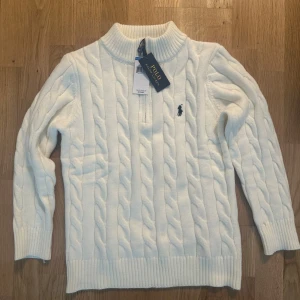 Vit kabelstickad tröja Polo Ralph Lauren - Vit kabelstickad  tröja från Polo Ralph Lauren med halv dragkedja och hög krage. Tröjan kan passa bra till S storlek också och den har lång ärm, ribbade muddar och klassisk broderad logga på bröstet. Perfekt för att ha på i vinter på någon mysig kväll, hör gärna av dig om du är intresserad av denna vara 