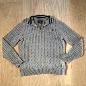 Grå stickad tröja från Polo Ralph Lauren - Säljer en grå kabelstickad tröja från Polo Ralph Lauren i storlek barn XL. Tröjan har halv dragkedja, ribbade muddar och krage med svarta och vita ränder.