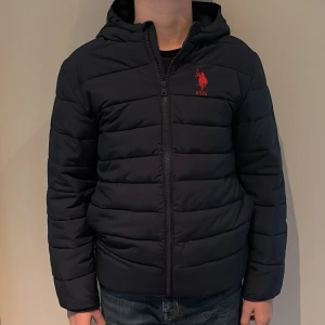 Svart pufferjacka från U.S.P.A - Mörkblå pufferjacka från U.S.P.A med röd broderad logga på bröstet. Jackan har huva, dragkedja framtill och quiltad design. Perfekt för kalla dagar.