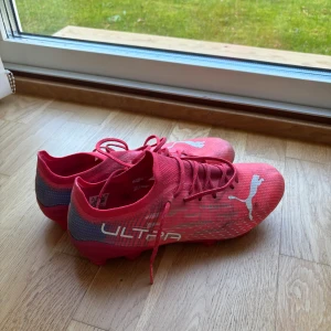 Röda Puma Ultra fotbollsskor - Säljer ett par röda Puma Ultra fotbollsskor med vita detaljer och snörning. Skorna har en smidig och lätt design med syntetmaterial och låg profil, perfekt för snabba rörelser på planen. Puma-loggan syns tydligt på ovansidan och sidan av skorna.
