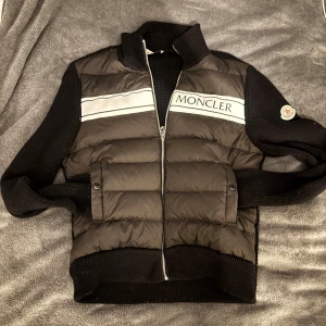 Moncler Cardigan (sällsynt) - Svart dunväst (cardigan) från Moncler med stickade svarta ärmar och ribbade muddar. Västen har en silverfärgad rand och Moncler-logga på bröstet samt dragkedja framtill. Klassisk quiltad look och diskret logotyp på ena ärmen. Perfekt för lager-på-lager-stil. Fungerande NFC