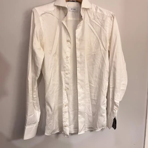 Vit skjorta från Eton, slim fit - Klassisk vit skjorta från Eton i slim fit med snyggt mönstrat tyg på insidan av kragen. Skjortan har långa ärmar, knappar framtill och är tillverkad i bomull. Perfekt för dig som gillar stilrena och fräscha plagg med en twist.