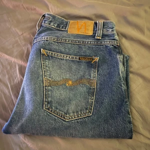 Blå jeans från Nudie Jeans, W32 L32 - Säljer ett par klassiska blå jeans från Nudie Jeans med snygga kontrastsömmar och ikonisk bakficka. Jeansen har rak passform och normal midja, tillverkade i slitstark bomull. Perfekta för dig som gillar en tidlös och avslappnad look.