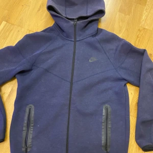 Mörkblå Nike hoodiejacka med dragkedja - Nike hoodiejacka i mörkblått med hel dragkedja och två fickor med svarta detaljer. Jackan har en sportig look, huva och diskret Nike-logga på bröstet. Tillverkad i mjukt material Den passar också folk som har S. 