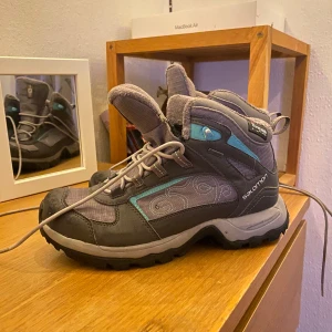 Gråa Salomon vinterkängor - Säljer ett par gråa Salomon vinterkängor med Thinsulate-foder och snygga turkosa detaljer. Kängorna har snörning, grov sula och är tillverkade i syntet och textil. Perfekta för kalla dagar och outdoor-äventyr. Mönstrad sida för extra stilpoäng.