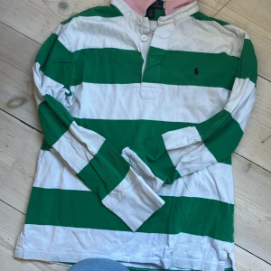 Randig pikétröja från Ralph Lauren, strl M 10-12 - Säljer en klassisk långärmad pikétröja från Ralph Lauren med breda gröna och vita ränder, rosa insida på kragen och broderad logga på bröstet. 