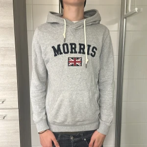 Grå hoodie från Morris med tryck - Snygg grå hoodie från Morris med broderad Union Jack-flagga och stort svart tryck på bröstet. Tröjan har huva med vita snören och en klassisk känguruficka framtill. Perfekt för chill dagar och enkel att matcha med jeans.