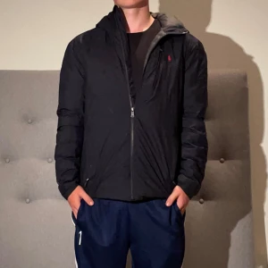 Mörkblå pufferjacka från Polo Ralph Lauren - Mörkblå pufferjacka från Polo Ralph Lauren med huva och dragkedja framtill. Jackan har diskret logga på bröstet och quiltad insida i grått. Perfekt för kyliga dagar och har en clean, sportig look. Jackan är i extremt bra skick!