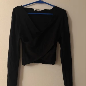 Svart omlottopp från Amisu - Svart långärmad omlottopp från Amisu med ribbad struktur. Toppen har en croppad passform och djup v-ringning framtill. Perfekt för dig som gillar stilrena och trendiga plagg med en twist.