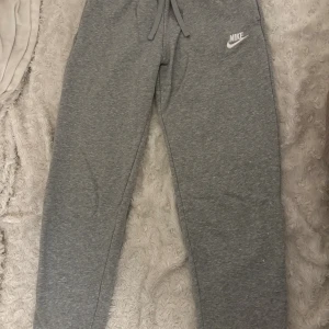 Grå mjukisbyxor från Nike XS - Grå mjukisbyxor från Nike i storlek XS med klassisk logga på låret. Byxorna har resår och snörning i midjan samt muddar vid bensluten. Tillverkade i mjukt bomullsmaterial för maximal komfort. Perfekta för chill eller träning. har små vita fläckar på vänstra ben.