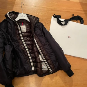 Moncler jacka med avtagbar huva - Säljer en mörkblå dunjacka från Moncler med glansigt, quiltat foder och huva. Luvan/inbyggda väst är avtagbar. Perfekt för kyliga dagar. Sitter som S
