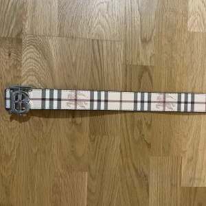 Burberry bälte med rutigt mönster - Säljer ett Burberry bälte med klassiskt rutigt mönster i beige, svart och rött på ena sidan och svart rutigt på andra. Bältet har en stor silvrig B-spänne och är gjort i skinn. Perfekt accessoar för att lyfta din outfit med en lyxig touch.