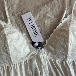 Vit spetstopp från Ivorie Studio - Supersöt vit topp från Ivorie Studio med tunna axelband, spetsdetaljer över bysten och en liten rosett framtill. Inlägg vid bysten. Aldrig använd med prislapp kvar. Nypris 639 kr