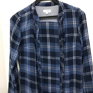 Blårutig skjorta med volang från KappAhl - Snygg blårutig skjorta från KappAhl i storlek 38. Skjortan har långa ärmar, knappar framtill och volangdetaljer längs knappraden. Perfekt för dig som gillar en avslappnad men ändå trendig look.