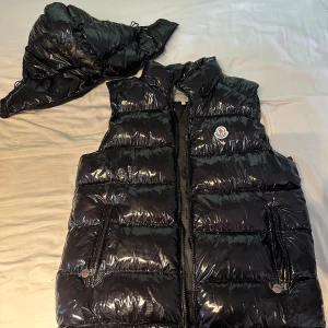 Svart dunväst från Moncler, storlek L - Säljer en NFC scanbar glansig svart dunväst från Moncler i storlek L. Västen har avtagbar huva, två dragkedjefickor framtill och klassisk Moncler-logga på bröstet. Perfekt för lager-på-lager och riktigt snygg till streetwear. Fodrad med dun och stängs med dragkedja.