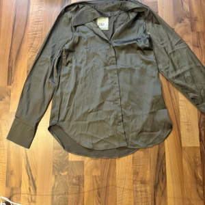 Olivgrön skjorta i satin, XS - Olivgrön skjorta i satinliknande material med klassisk krage och knäppning framtill. Skjortan har långa ärmar med breda manschetter och en lätt glansig finish som ger en stilren vibe. Perfekt för dig som gillar en clean och enkel look.