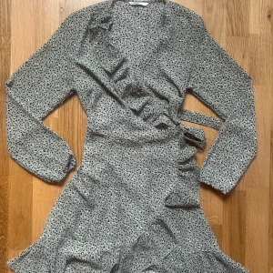 Prickig omlottklänning från ONLY - Superfin kort omlottklänning från ONLY i ljusgrått med svarta prickar. Klänningen har långa ärmar, volangdetaljer längs v-ringningen och knyt i midjan. Perfekt för dig som gillar en lekfull och trendig stil.