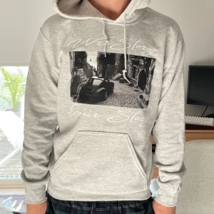 Grå Limiterad WriStory Hoodie - Snygg ljusgrå hoodie från WriStory med svartvitt tryck av en bil och texten 'Your Story' på bröstet. Tröjan har huva med snörning, känguruficka och mjukt material i bomullsblandning. Perfekt för chill dagar eller när du vill ha en avslappnad streetstyle. 