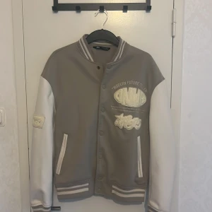 Grå och vit bomberjacka från Zara - Snygg bomberjacka i grått och vitt från Zara med broderade patches och textdetaljer som 'Modern Future Teller' och '1975'. Jackan har ribbade muddar, tryckknappar och två fickor framtill. Perfekt för dig som gillar streetstyle och coola detaljer.