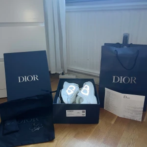 Vita Dior B30’s sneakers med reflekterande detaljer - Säljer ett par exklusiva vita Dior sneakers med reflekterande silverdetaljer och tydlig Dior-logga på sidan. Skorna har snörning, rund tå och platt sula. Kommer med originalkartong, dustbag och shoppingpåse. Perfekta för dig som vill sticka ut med lyxig streetstyle.