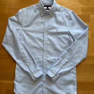 Ljusblå skjorta Tommy Hilfiger slim fit - Snygg ljusblå skjorta från Tommy Hilfiger i slim fit-modell. Skjortan har klassisk krage, knäppning framtill och broderad logga på bröstet. Tillverkad i bomull och har långa ärmar med knappar vid manschetterna.