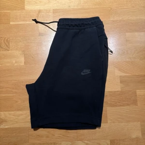 Svarta Nike tech fleece shorts - Svarta shorts från Nike med elastisk midja och snörning. Snygg minimalistisk look med diskret Nike-logga på ena benet och praktisk ficka med dragkedja på sidan. Perfekta för chill eller träning, tillverkade i mjuk bomullsmix.