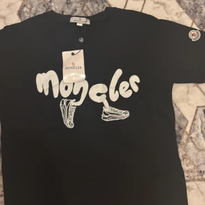Svart Moncler t-shirt med tryck - Svart t-shirt från Moncler med coolt vitt tryck framtill där loggan är stylad med sneakers. Klassisk rund hals och korta ärmar. Moncler-märke på ärmen. Perfekt för dig som gillar streetstyle och vill sticka ut. Priset kan diskuteras!!