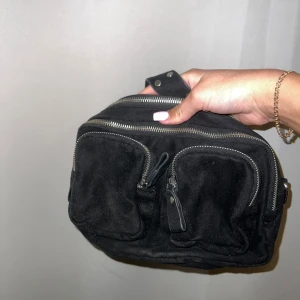 Svart axelväska i mocka med nitar - Cool svart axelväska i mocka med bred axelrem dekorerad med silvriga nitar. Väskan har flera fack med dragkedjor och två praktiska ytterfickor framtill. Följer med ett längre band, från Gina tricot