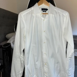 Vit skjorta från Riley i bomull - Klassisk vit skjorta från Riley med lång ärm och snygg krage. Skjortan har knappar framtill och är tillverkad i mjuk bomull. Perfekt för dig som gillar en clean och stilren look. Använts enbart fåtal gånge och sedan legat i garderoben. Bekväm o fint material. 