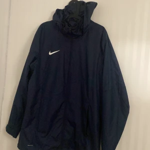 Mörkblå Nike vind/regnjacka med huva  - Snygg mörkblå vindjacka från Nike med huva och dragkedja framtill. Jackan har Nike-logga på bröstet och är tillverkad i lätt polyester. Perfekt för blåsiga eller regniga dagar och sportiga outfits. Modellen är enkel och stilren med långa ärmar och praktiska fickor. Köpt på Intersport för något år sen.